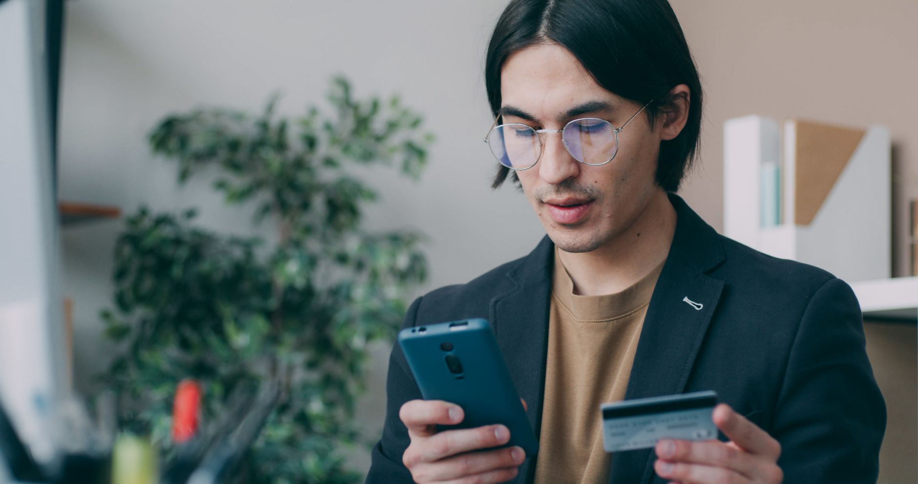 Homem jovem com óculos, segurando celular e cartão, em ambiente interno, olhando para a tela.